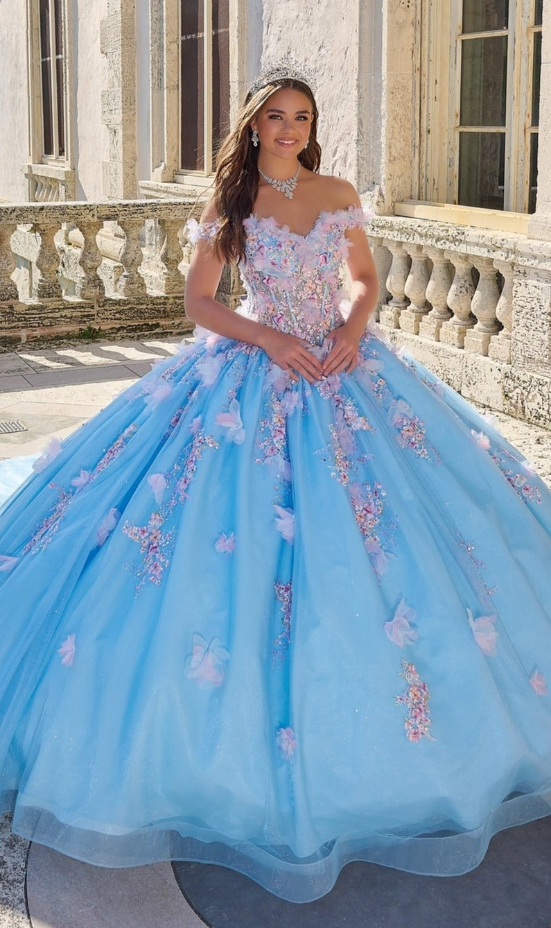 A young woman wearing the Quinceanera Dress 54228 By Amarra with delicate 3D floral appliqué stands outdoors in front of a stone balustrade and building.